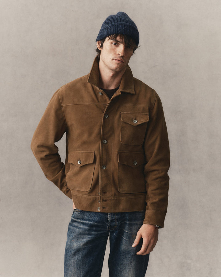 A man wearing a brown suede jacket, blue jeans, and a navy blue knit beanie, standing against a textured gray wall.