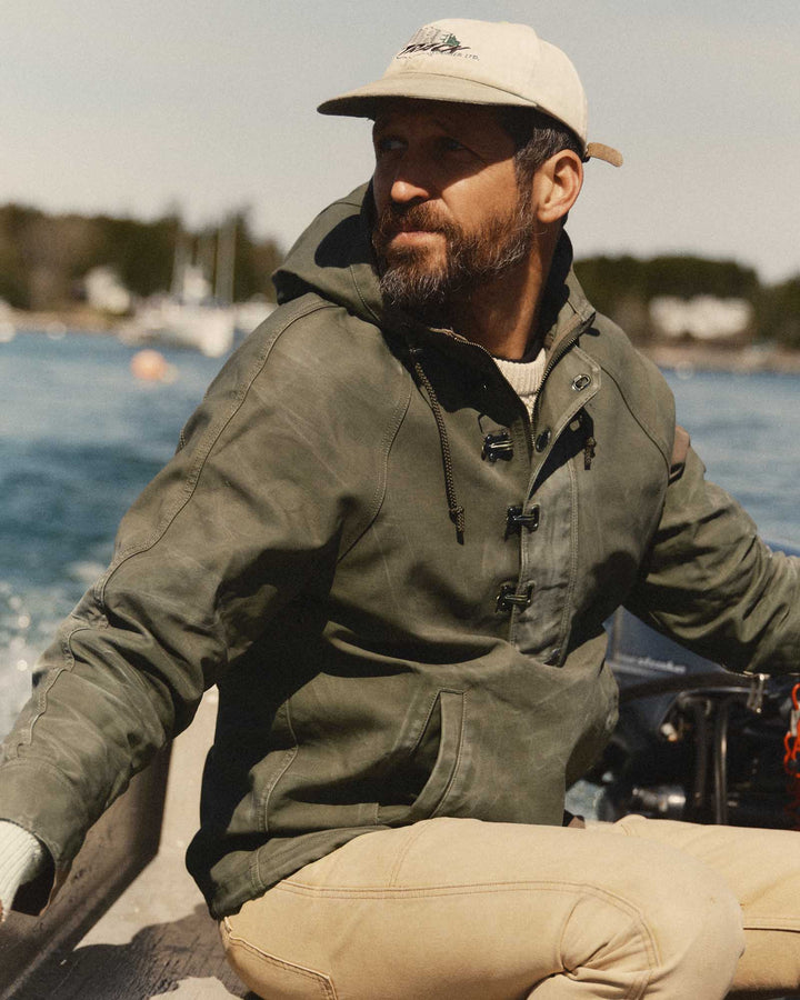 A man wearing an olive green hooded hatteras anorak jacket, beige pants, and a beige baseball cap sits on a boat with water and a shoreline in the background.