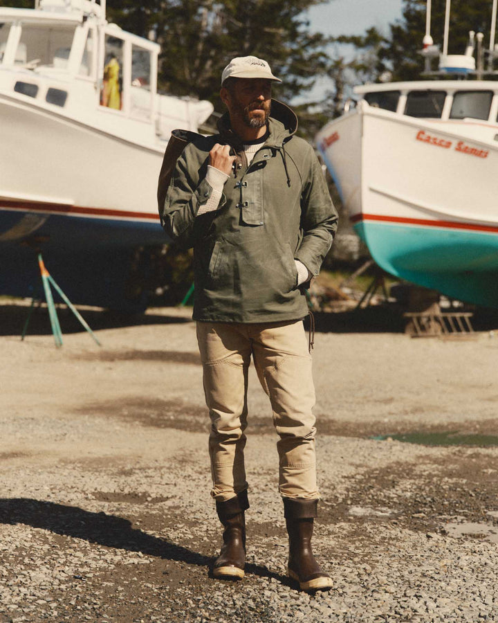 A man stands outdoors in a boatyard wearing an olive green hooded Hatteras anorak jacket, beige pants, and dark brown rubber boots. White boats are parked in the background on a gravel ground.