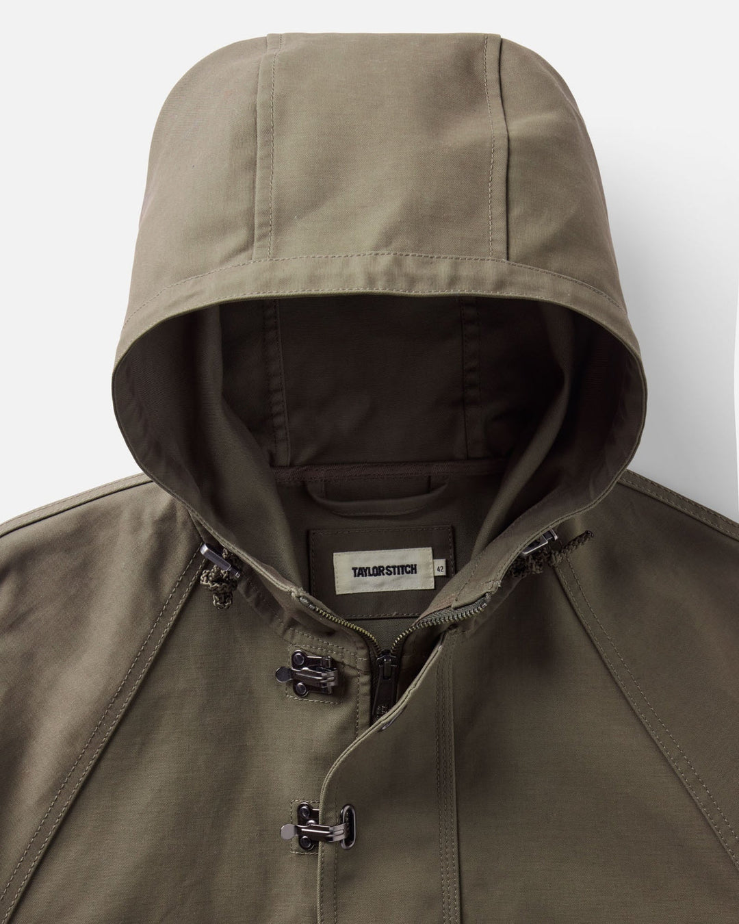 Close-up of an olive green hooded Hatteras anorak jacket with a front zipper, black toggle closures, and a visible Taylor Stitch brand label inside the collar.