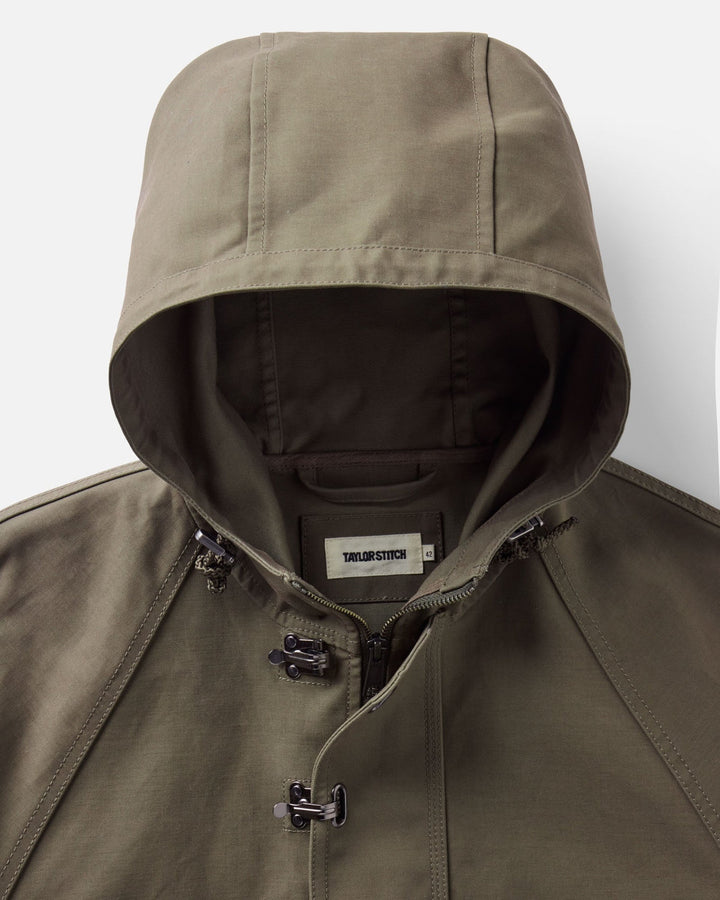 Close-up of an olive green hooded Hatteras anorak jacket with a front zipper, black toggle closures, and a visible Taylor Stitch brand label inside the collar.