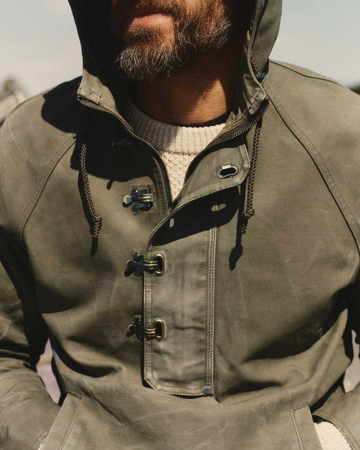 Close-up of a man wearing an olive green Hatteras anorak jacket with toggle closures and a cream knit sweater underneath, photographed outdoors in natural light.
