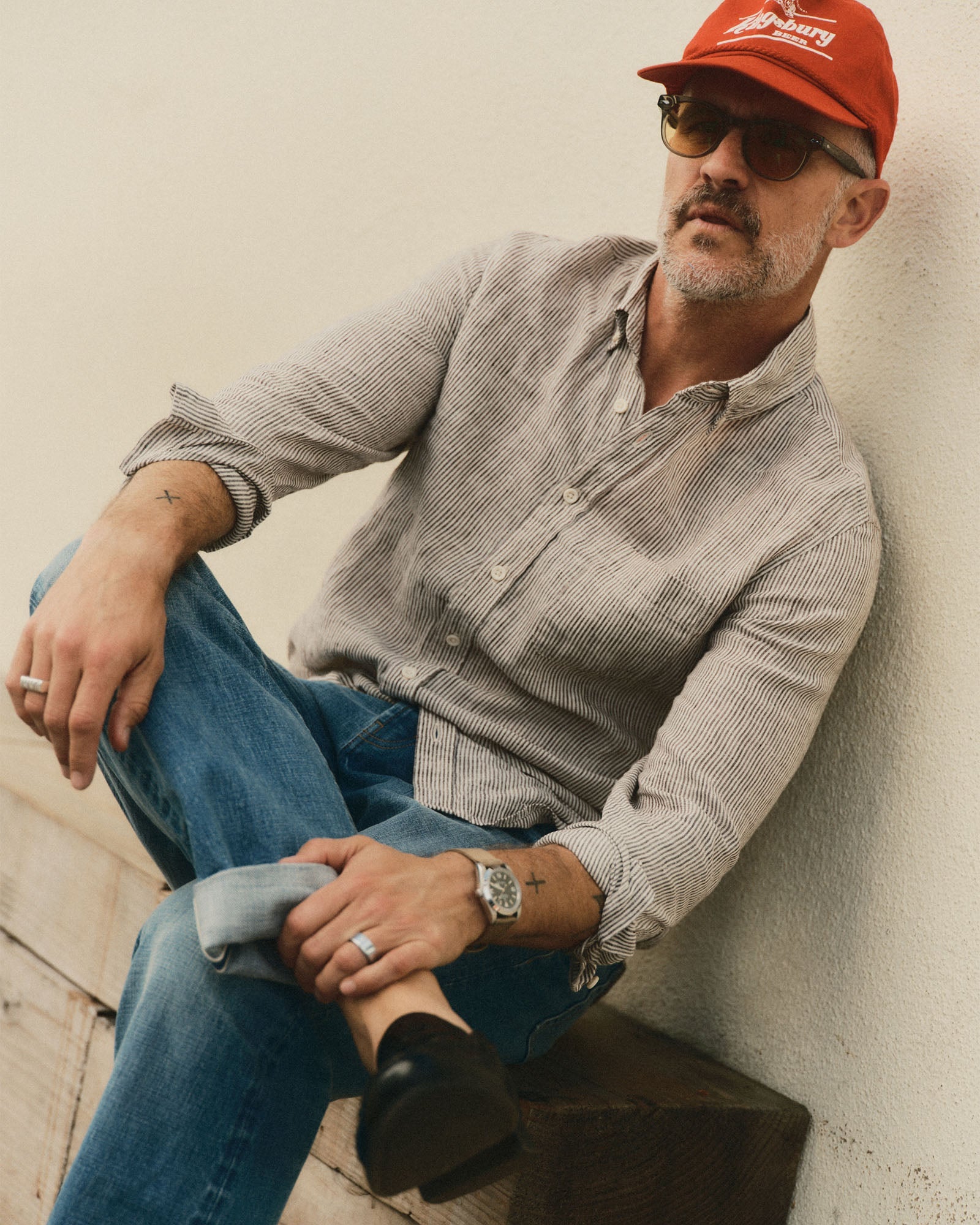 Man wearing a red cap, sunglasses, jeans and the jack shirt in burgundy university stripe linen leaning against a wall.