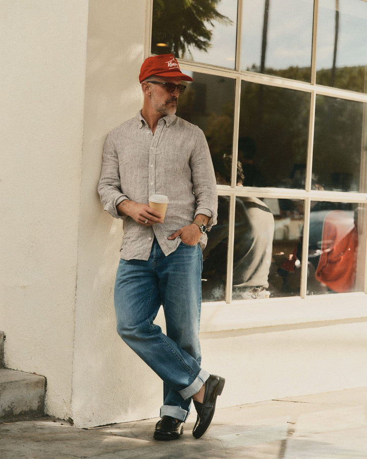Man wearing a red cap, sunglasses, jeans and the jack shirt in burgundy university stripe linen leaning against a building.