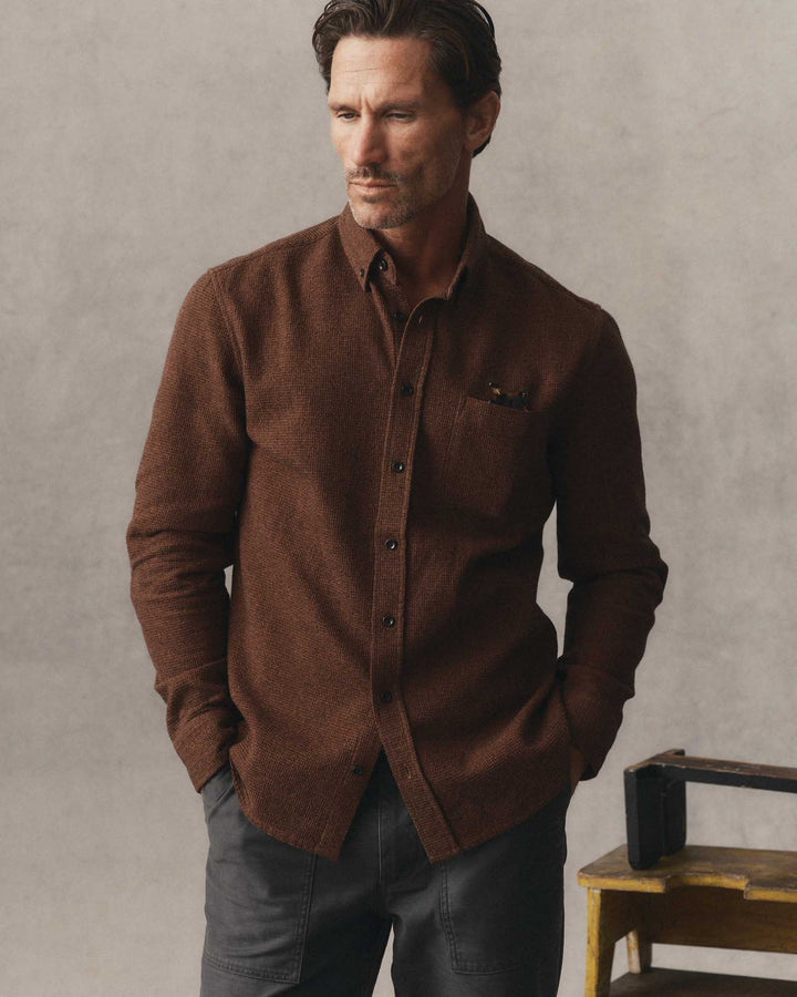 A man wearing The Jack in Timber Twist Waffle and dark gray trousers, standing against a textured light gray wall with a small wooden stool visible on the right.