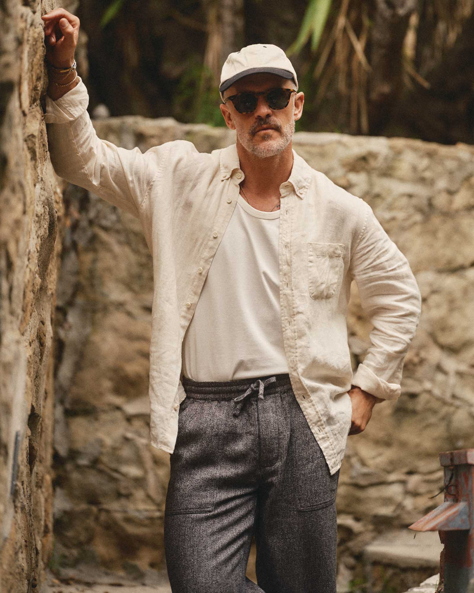 Man wearing sunglasses, a cap, and a light-colored shirt leaning against a stone wall.