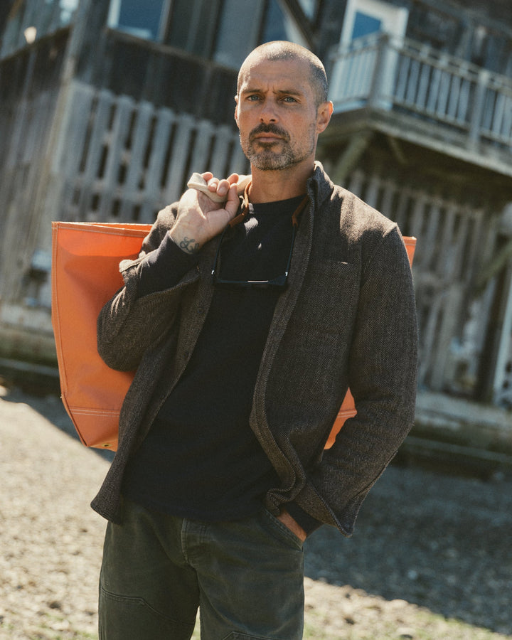 A man wearing a brown jack shirt over a black shirt and dark pants, holding an orange tote bag against a rustic wooden building background.