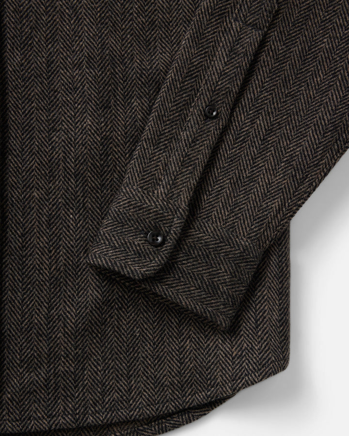 Close-up of a dark brown and black herringbone patterned fabric, showing a folded cuff with black buttons on a light gray background.