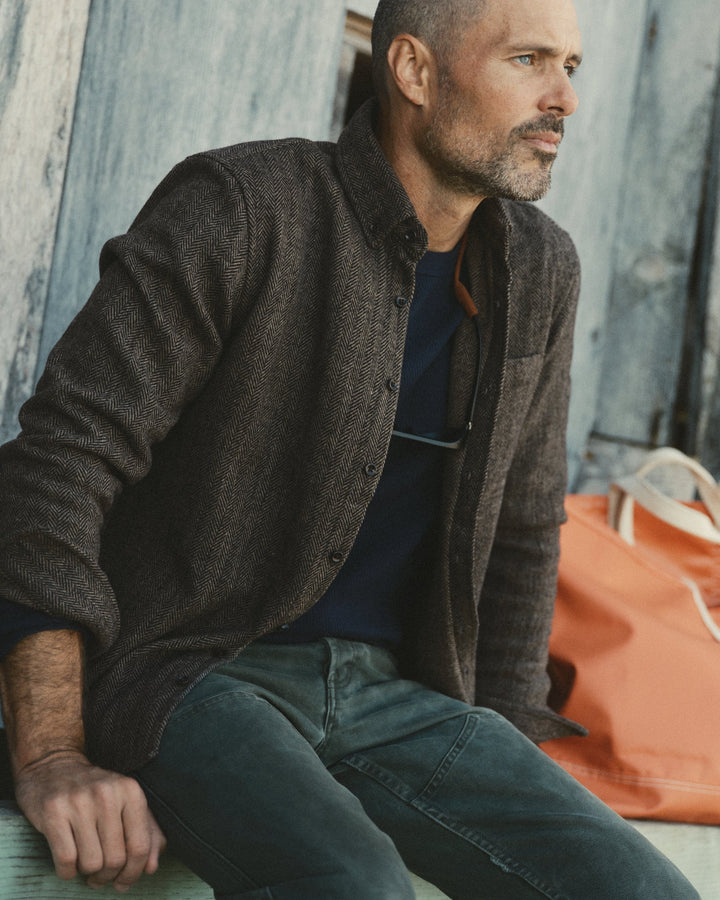 A man wearing a dark brown textured button-up overshirt over a navy t-shirt and blue jeans, sitting against a weathered blue wooden wall with an orange tote bag beside him.