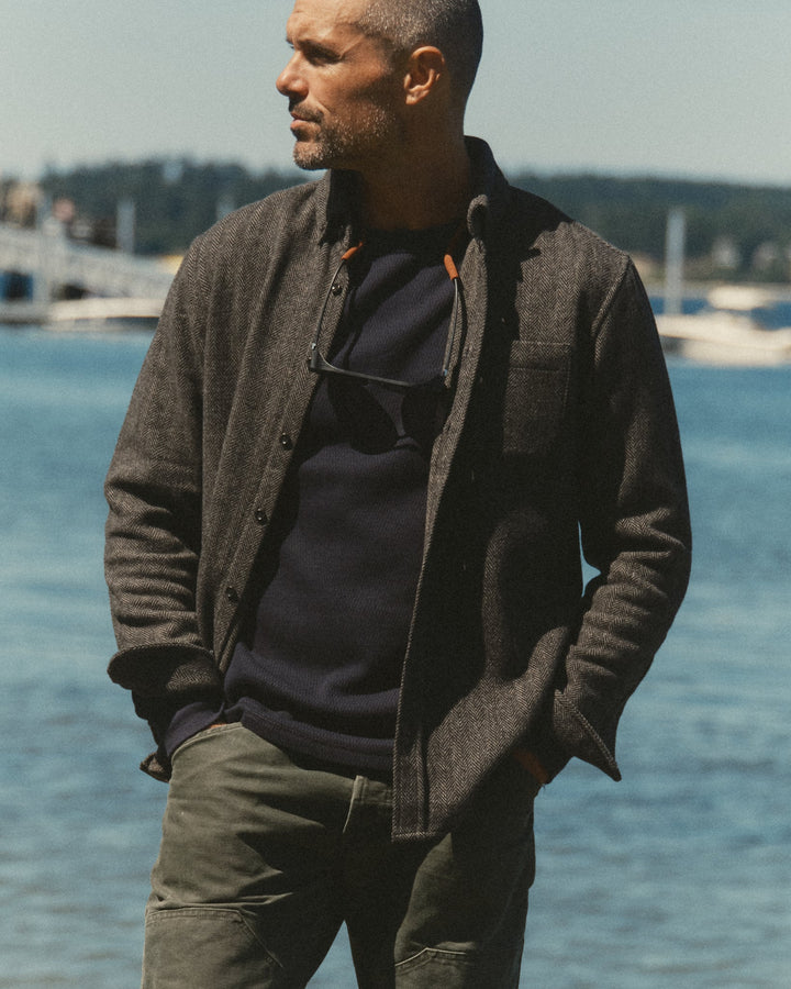 A man standing outdoors by the water wearing a dark charcoal wool-blend button-front overshirt over a navy sweater and olive pants.