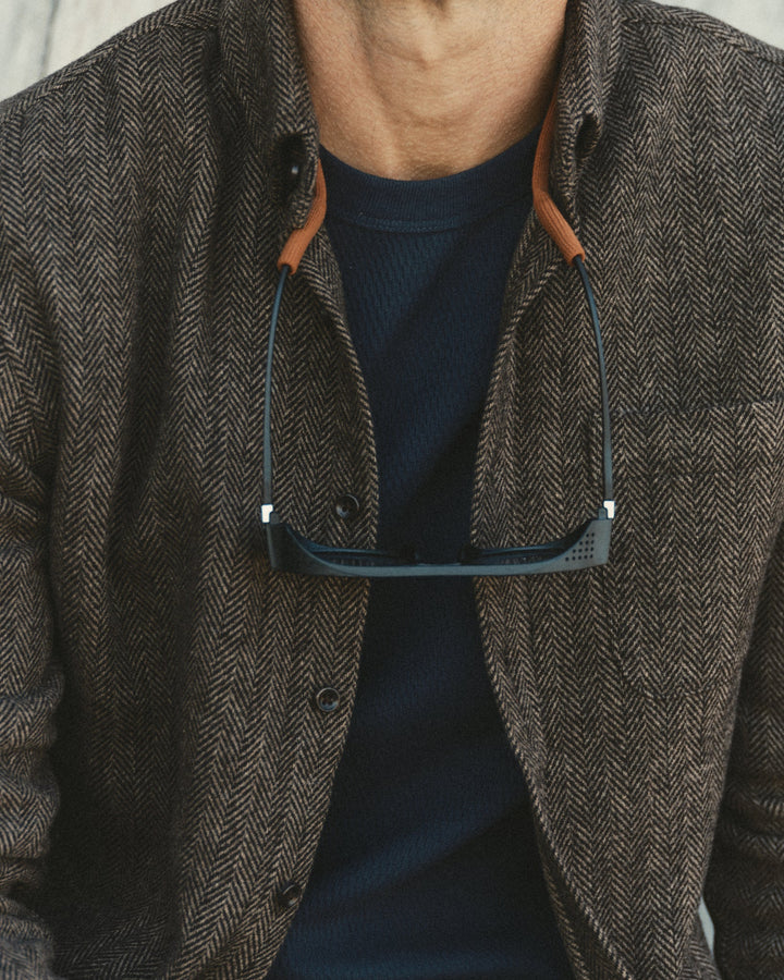 Close-up of a person wearing a brown herringbone jacket over a navy ribbed t-shirt, with dark-framed sunglasses hanging from the collar.