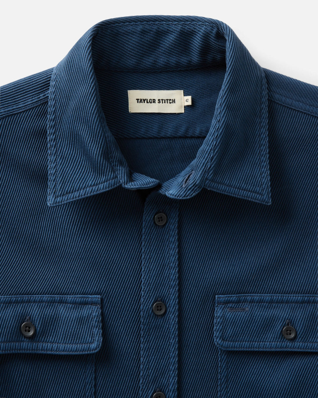 Close-up of a navy blue textured button-down shirt by Taylor Stitch, showing the collar, front buttons, and chest pockets against a light background.