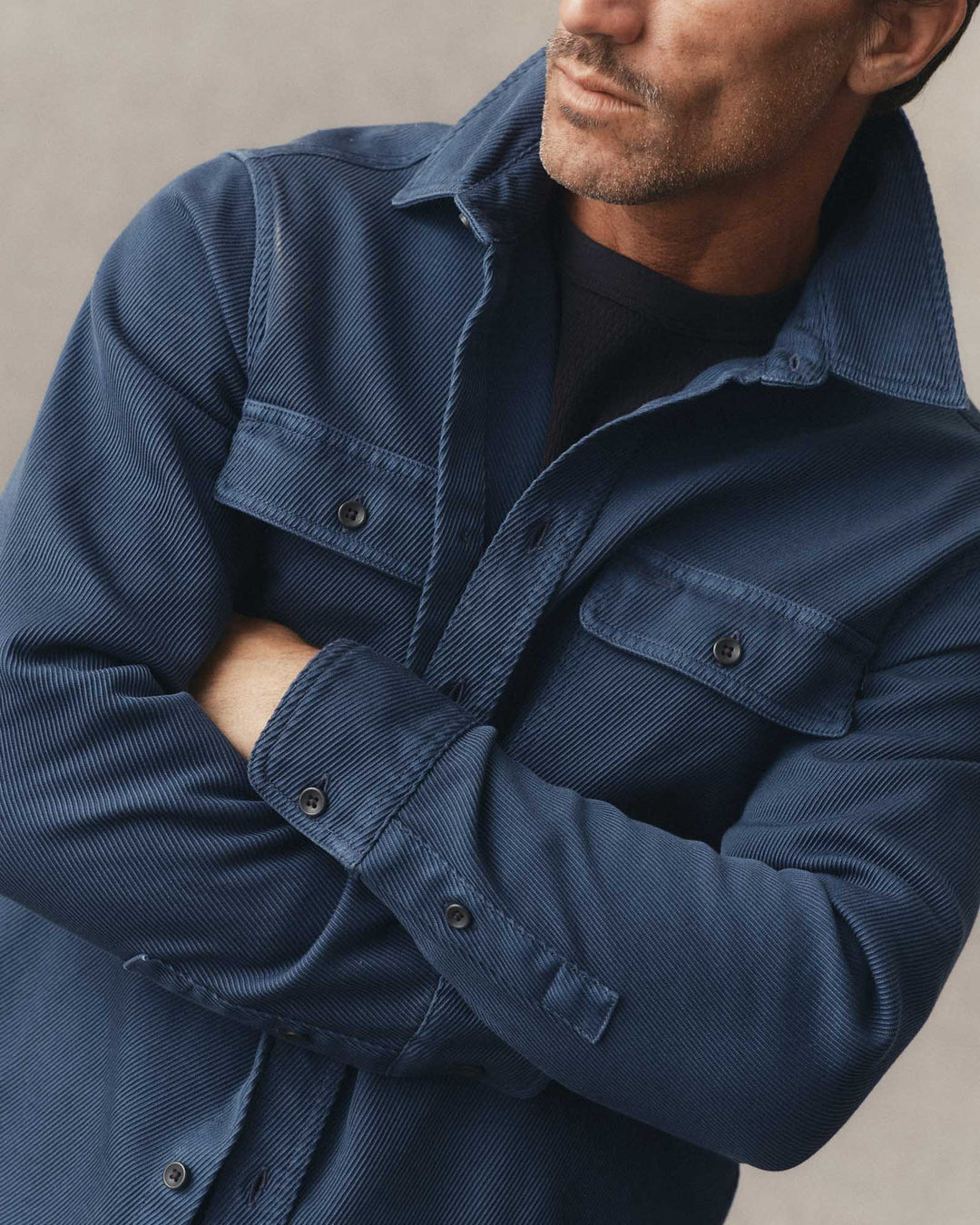 A close-up of a man wearing a textured navy blue corduroy overshirt over a black t-shirt, with his arms crossed.
