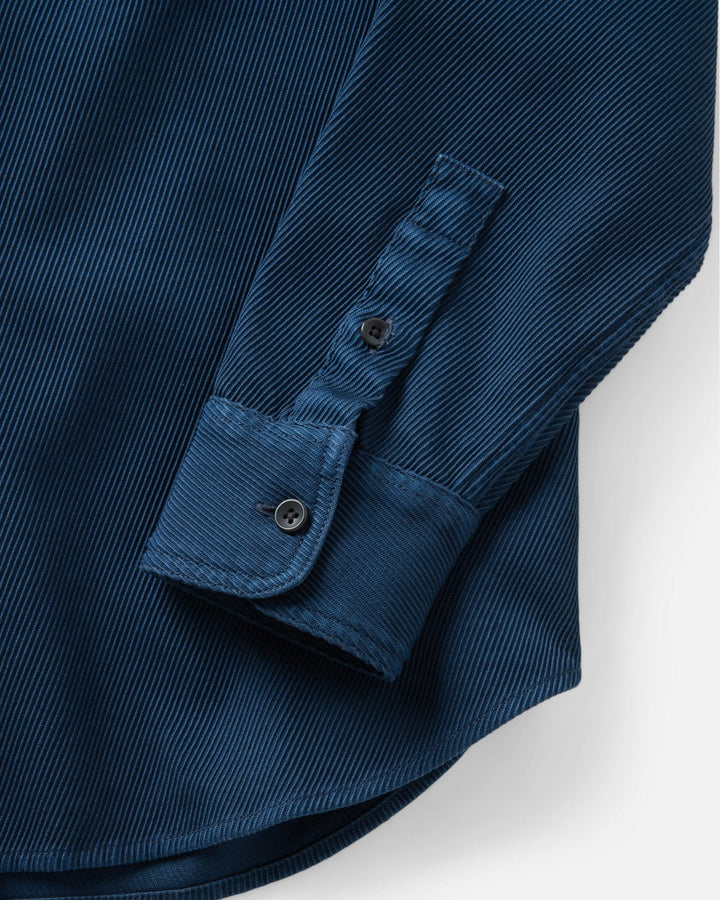 Close-up of a dark blue corduroy shirt sleeve with a buttoned cuff, showing the ribbed texture against a white background.