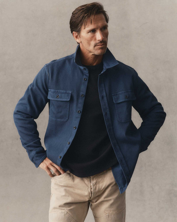 A man wearing a navy blue button-up overshirt jacket over a black t-shirt and beige pants, standing against a textured gray wall.