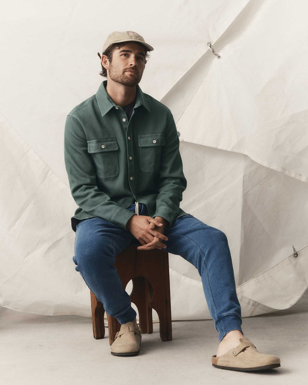A man sits on a wooden stool wearing a deep sea ledge button-up shirt, blue jeans, beige slip-on shoes, and a beige cap, posed against a draped white fabric backdrop.