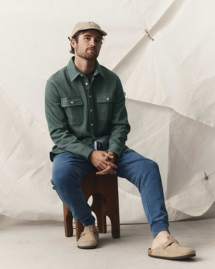 A man sits on a wooden stool wearing a deep sea ledge button-up shirt, blue jeans, beige slip-on shoes, and a beige cap, posed against a draped white fabric backdrop.