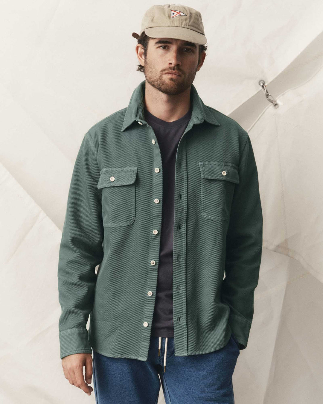 A man wearing a ledge deep sea button-up shirt jacket over a dark t-shirt and blue jeans, with a beige cap, posed against a white tent-like backdrop.