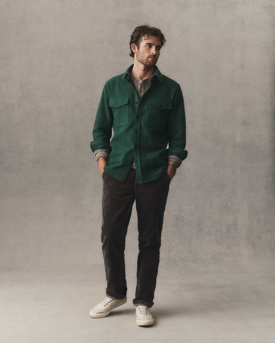 A man wearing a dark forest button-up maritime shirt jacket over a gray undershirt, paired with dark brown pants and white sneakers, standing against a textured gray studio backdrop.
