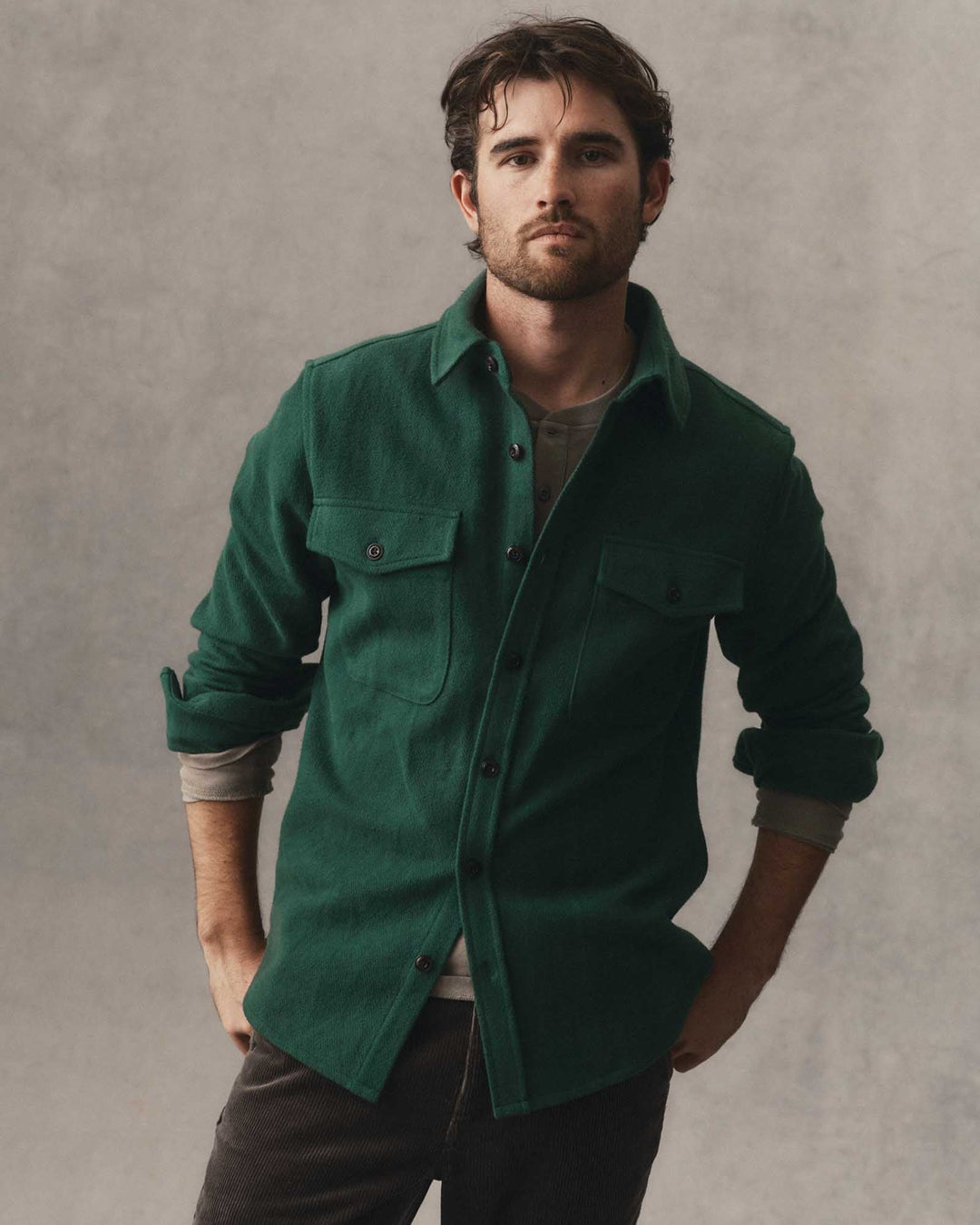A man wearing a dark forest maritime shirt jacket over a gray t-shirt and dark pants, standing against a textured gray studio background.