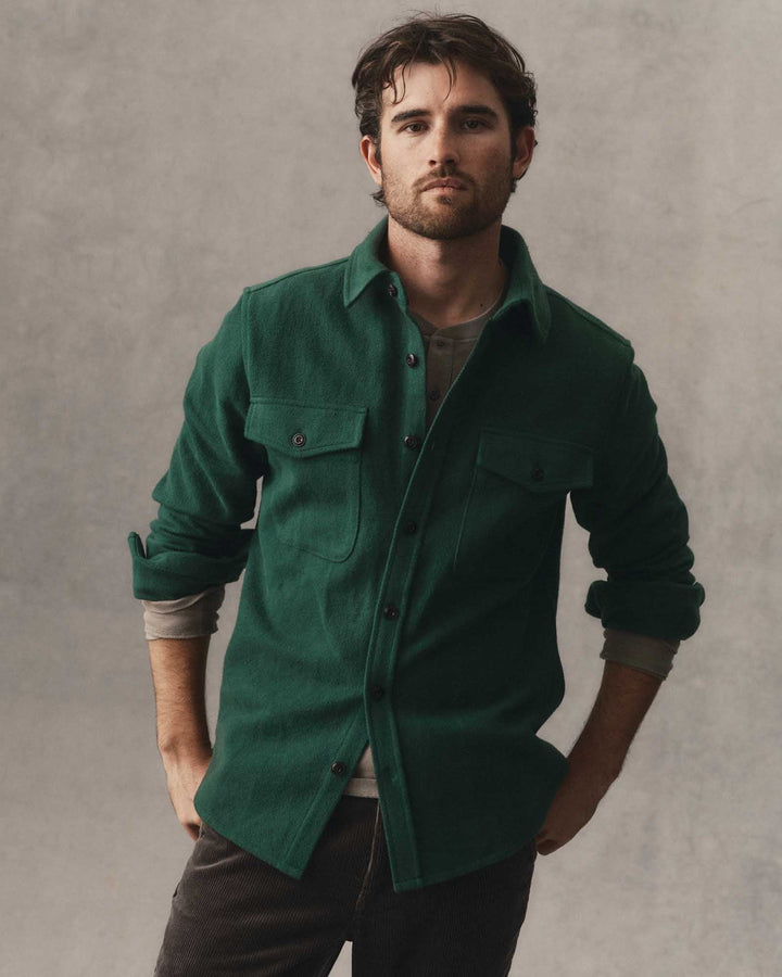 A man wearing a dark forest maritime shirt jacket over a gray t-shirt and dark pants, standing against a textured gray studio background.
