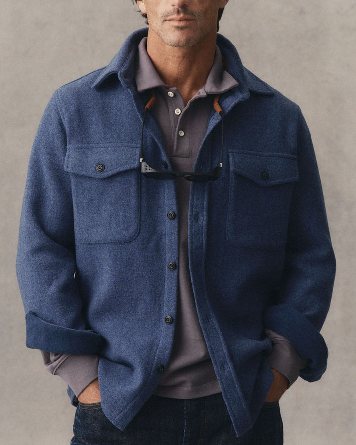 A man wearing The Maritime Shirt Jacket
in Heather Navy Wool over a gray polo shirt, with sunglasses hanging from the collar, set against a textured gray background.