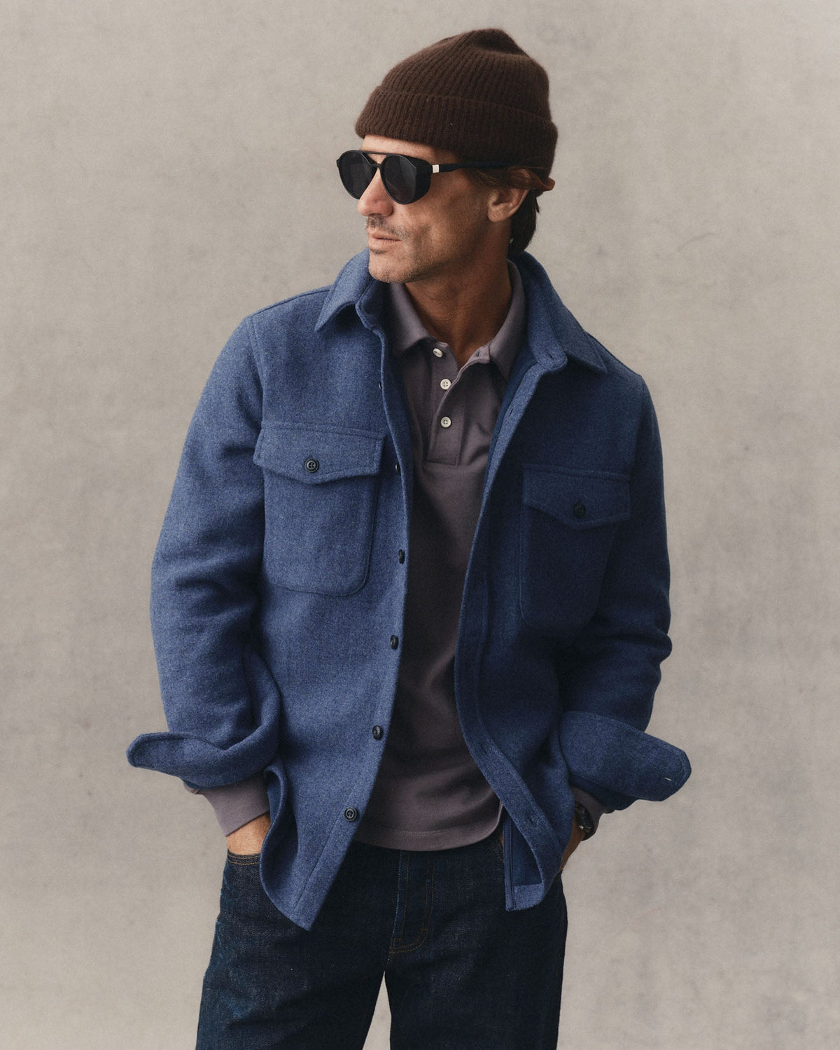 A man wearing The Maritime Shirt Jacket
in Heather Navy Wool, dark jeans, a brown knit beanie, and sunglasses, standing against a textured gray wall.