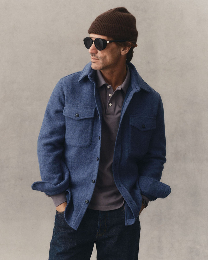 A man wearing The Maritime Shirt Jacket
in Heather Navy Wool, dark jeans, a brown knit beanie, and sunglasses, standing against a textured gray wall.