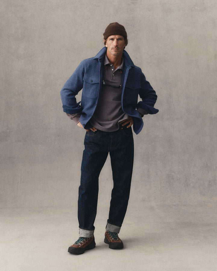 A man stands against a textured gray studio backdrop wearing The Maritime Shirt Jacket in Heather Navy Woolover a gray polo shirt, dark blue jeans, and brown lace-up shoes with a brown beanie.