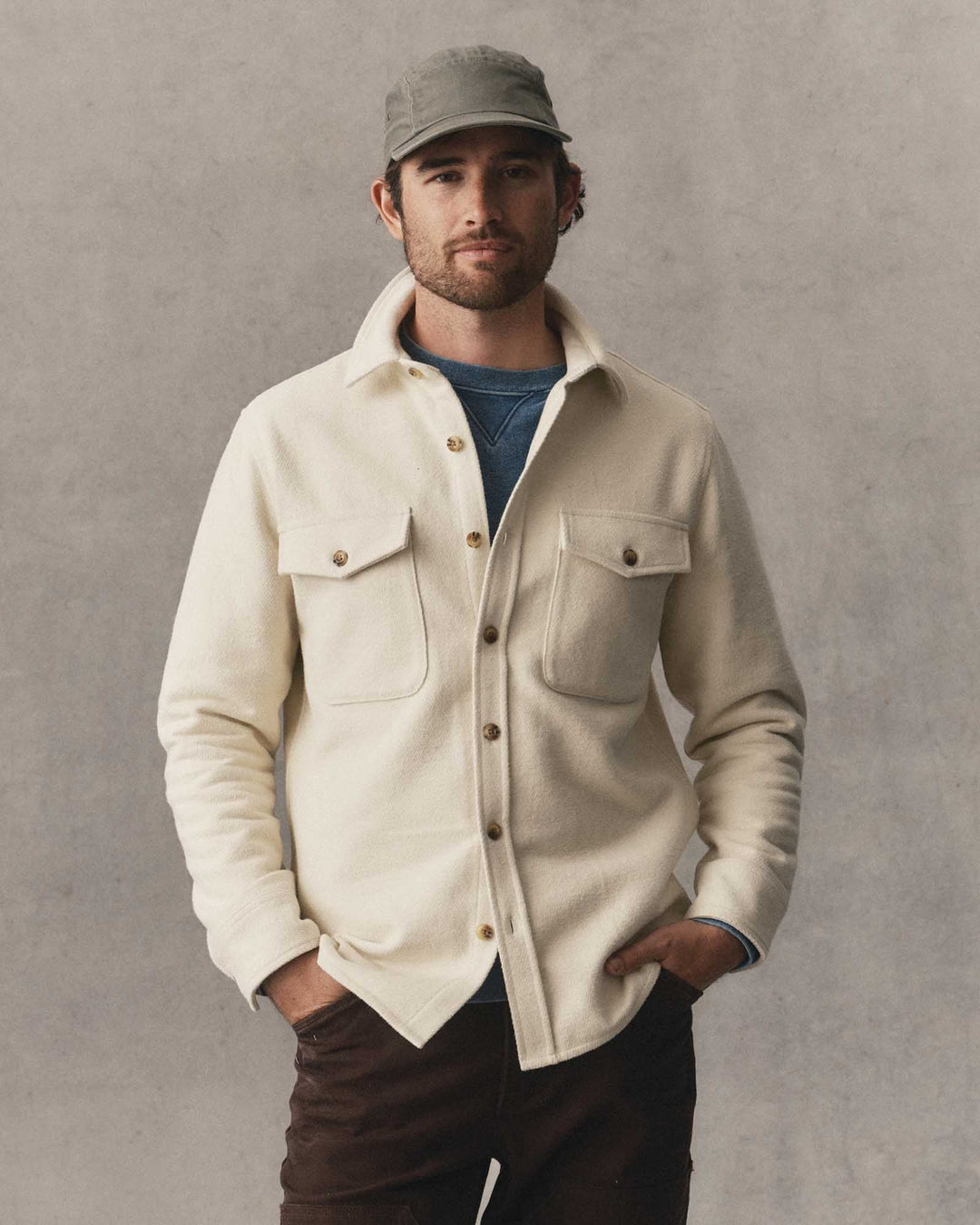 A man wearing a vintage white-colored button-up maritime shirt jacket over a blue t-shirt, paired with dark brown pants and a gray cap, standing against a textured gray background.