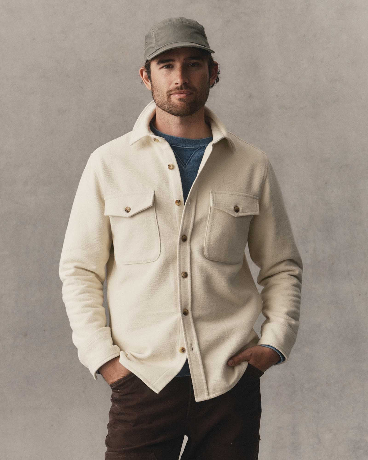 A man wearing a vintage white-colored button-up maritime shirt jacket over a blue t-shirt, paired with dark brown pants and a gray cap, standing against a textured gray background.
