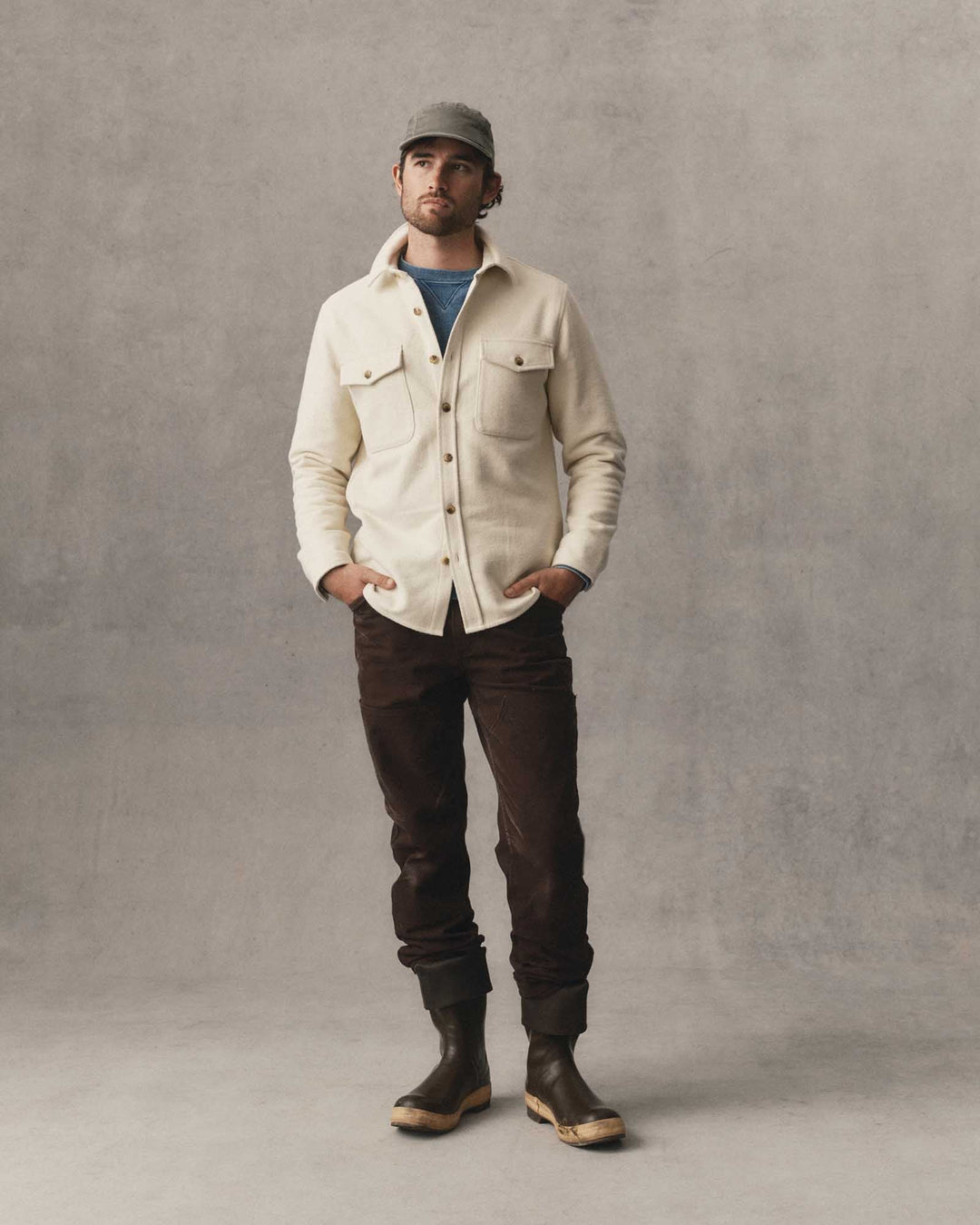 A man stands against a textured gray studio backdrop wearing a vintage white-colored button-up overshirt over a blue t-shirt, dark brown pants, and dark brown rubber boots.