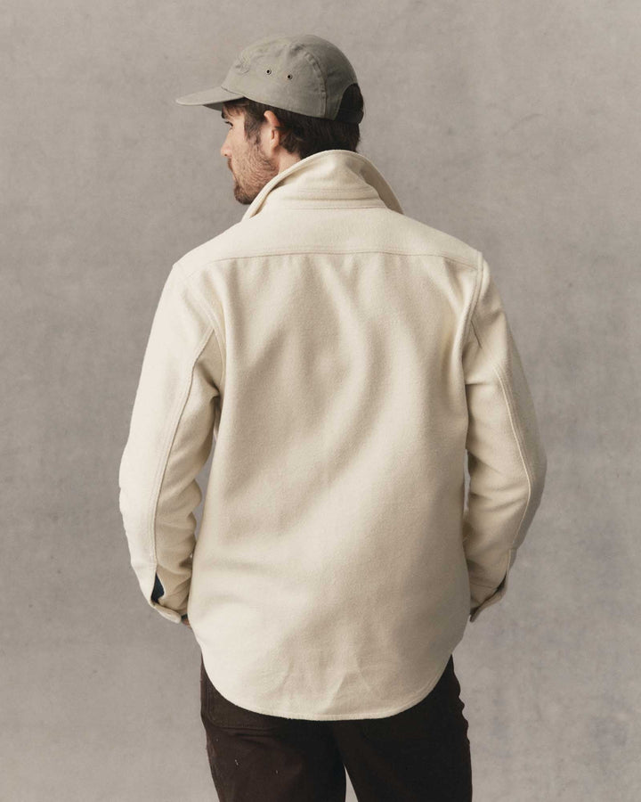 A man wearing a vintage white maritime shirt jacket and a gray baseball cap, photographed from behind against a textured gray wall.