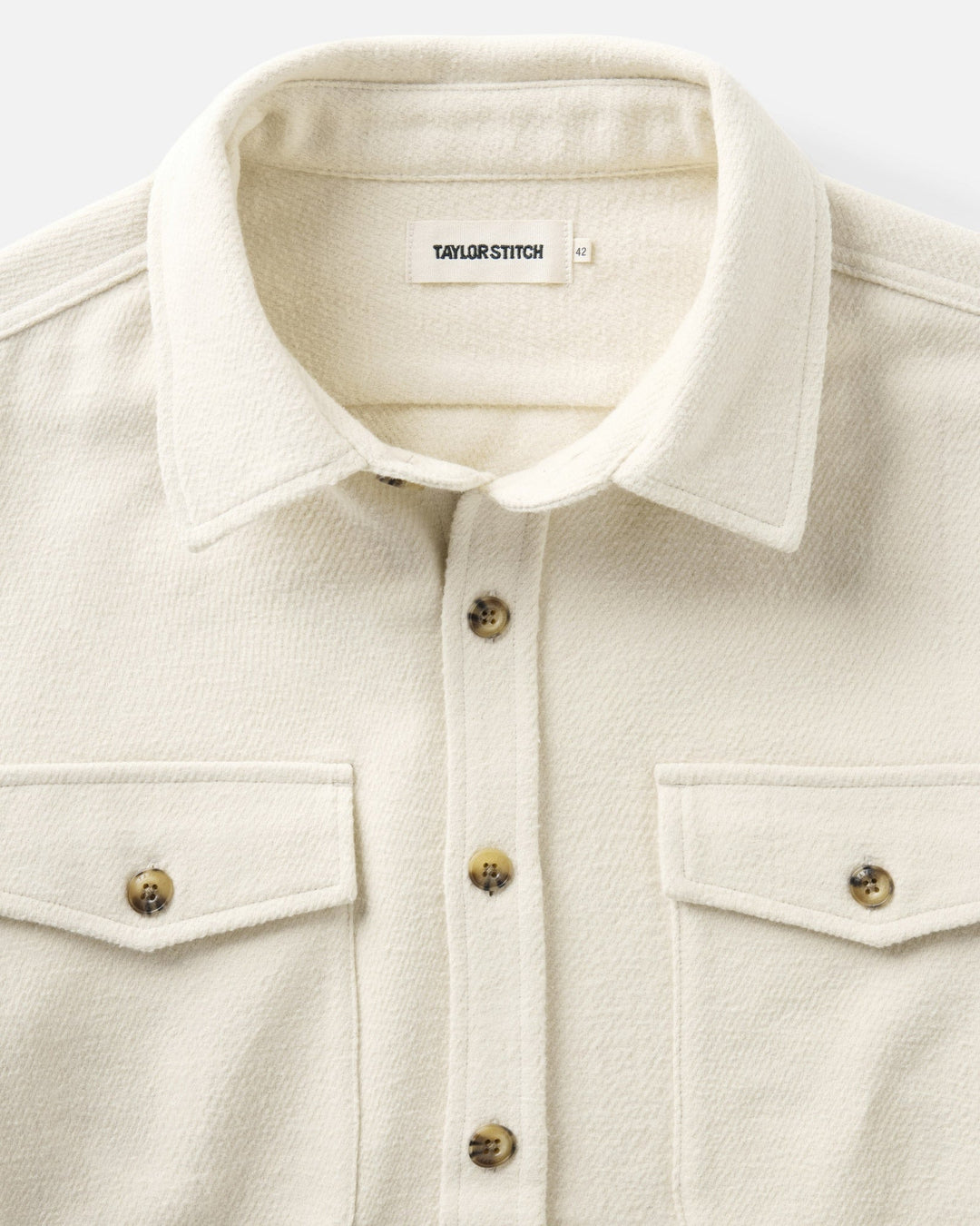 Close-up of a vintage white-colored maritime shirt jacket by Taylor Stitch, featuring a pointed collar, buttoned front, and two chest flap pockets with decorative brass-tone buttons.