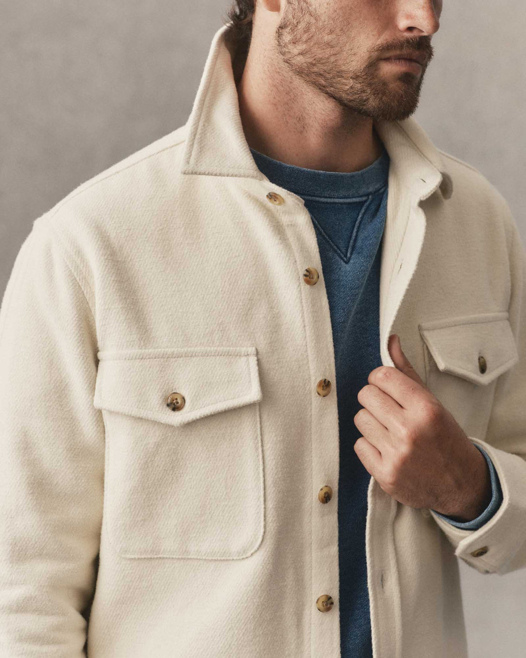 A close-up of a man wearing a vintage white-colored maritime shirt jacket over a blue crewneck sweater, set against a neutral gray background.
