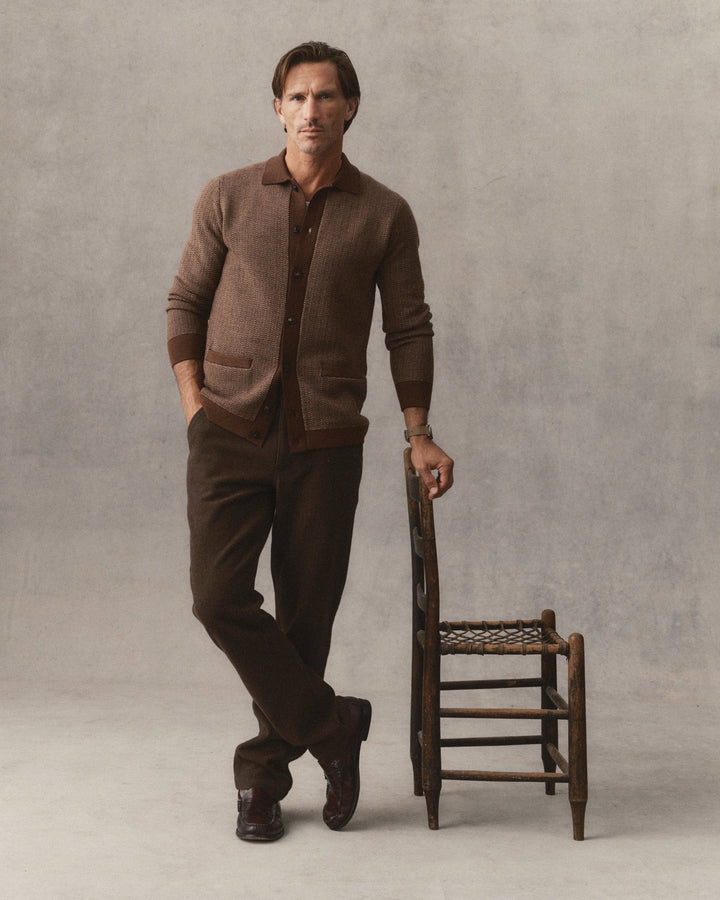A man stands in a studio wearing The Miles Sweater Polo in Espresso Merino over a collared shirt and dark brown trousers, leaning against a small wooden chair against a textured gray backdrop.