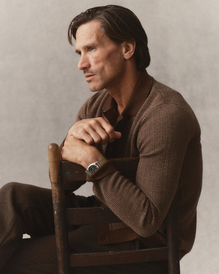 A man seated on a wooden chair wearing The Miles Sweater Polo in Espresso Merino over a dark brown shirt and brown trousers, photographed against a neutral light gray studio background.