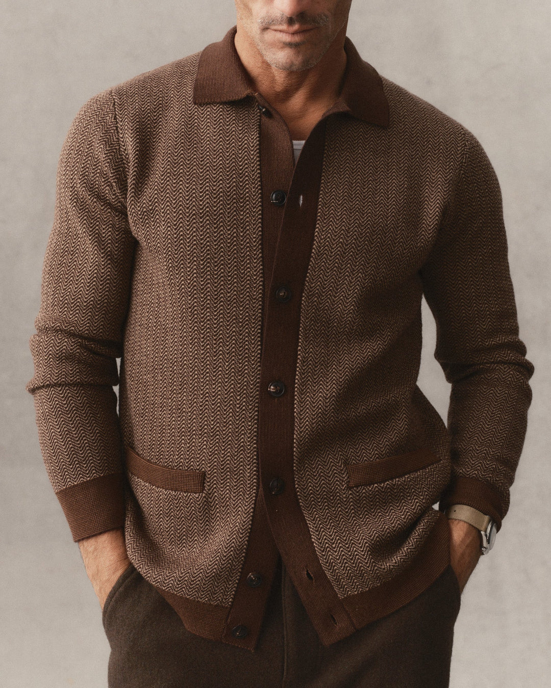 A man wearing The Miles Sweater Polo
in Espresso Merino with solid brown collar, cuffs, and pockets, paired with dark brown trousers against a neutral gray background.