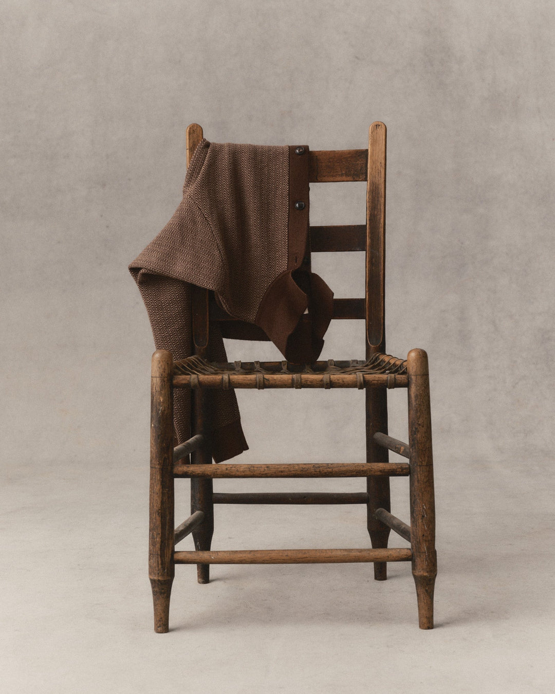 The Miles Sweater Polo in Espresso Merino draped over a rustic wooden chair with a woven seat, set against a neutral gray studio backdrop.