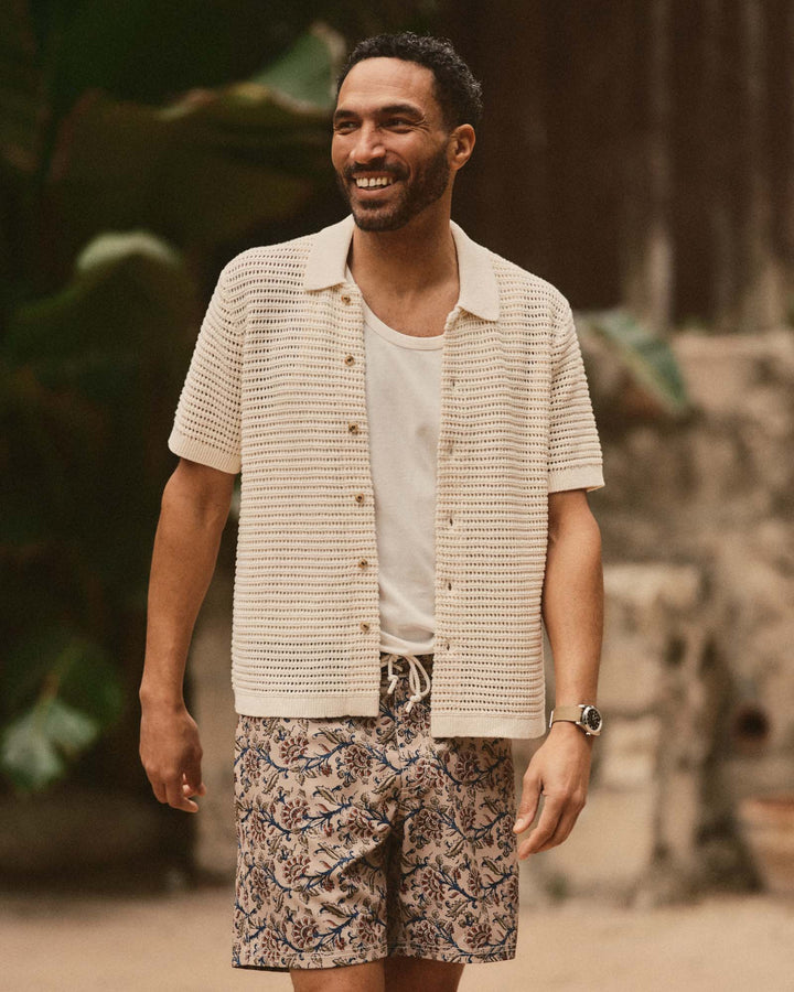 Man wearing The Monterey Sweater Polo in Vintage White Crochet and patterned shorts standing outdoors with greenery in the background