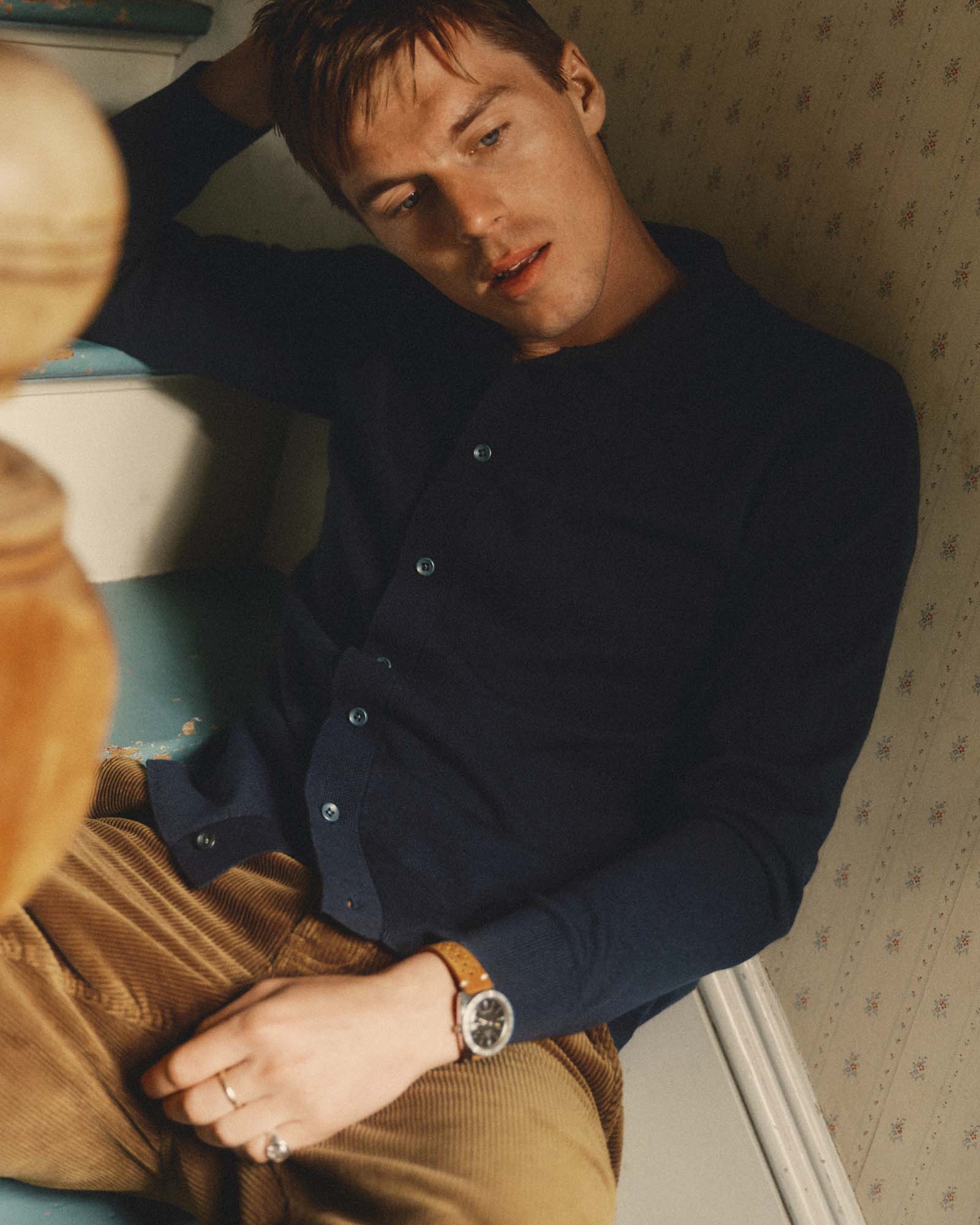 A man wearing Taylor Stitch's The Nichols Polo in Nocturne Cotton Cashmere and brown corduroy trousers, leaning against a patterned wall. He is wearing a brown leather strap watch on his wrist.