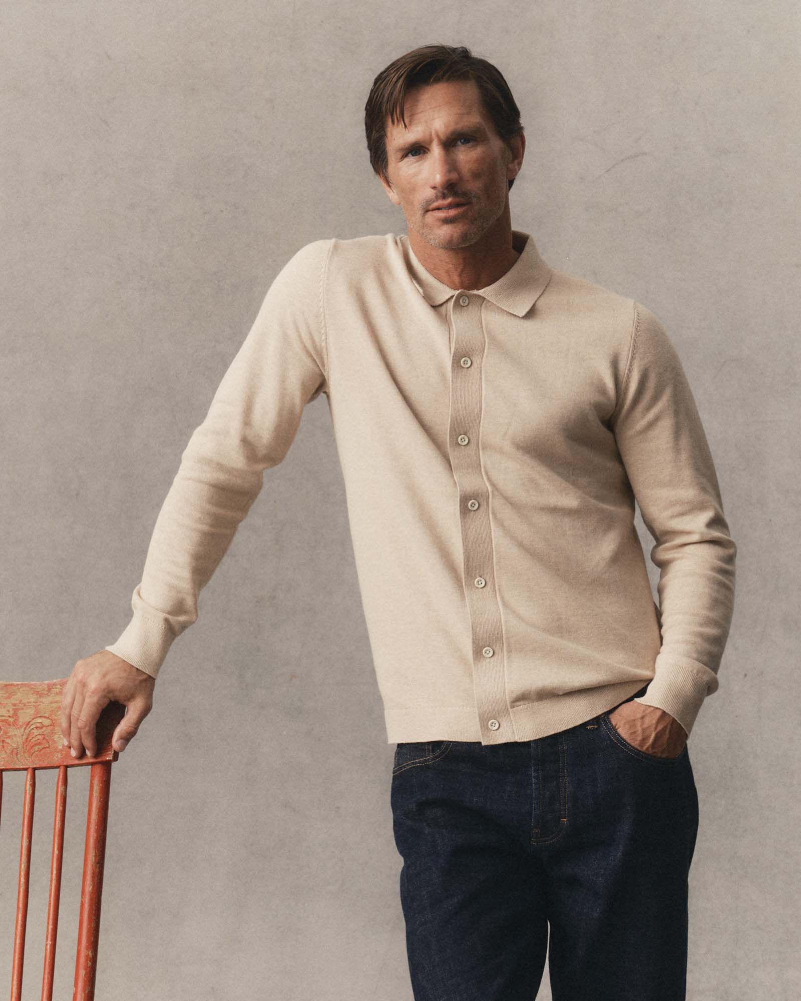 A man wearing Taylor Stitch's Nichols Polo Sable Cotton Cashmere and dark blue jeans, leaning against a wooden chair against a textured gray wall.