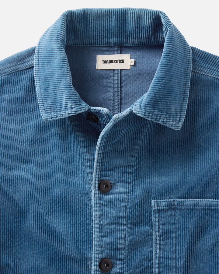 Close-up of a washed indigo corduroy button-up ojai jacket with a pointed collar and dark buttons, showing the Taylor Stitch brand label inside the neck.