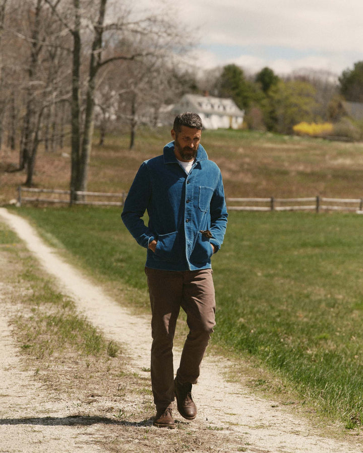 A man walking on a dirt path in a rural outdoor setting, wearing a washed indigo corduroy ojai jacket, brown pants, and brown boots.