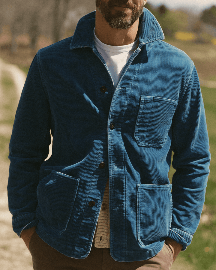 A man wearing a washed indigo corduroy Ojai button-up jacket over a white t-shirt and beige sweater, standing outdoors in a grassy field.