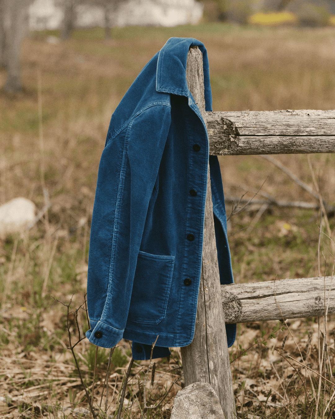 A washed indigo corduroy ojai jacket hanging on a weathered wooden fence post in an outdoor field.