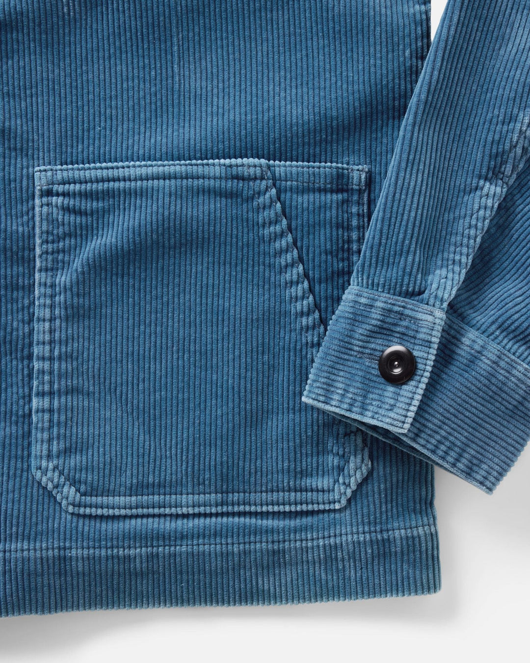 Close-up of a washed indigo corduroy ojai jacket showing a front patch pocket and the folded cuff of a long sleeve with a black button.