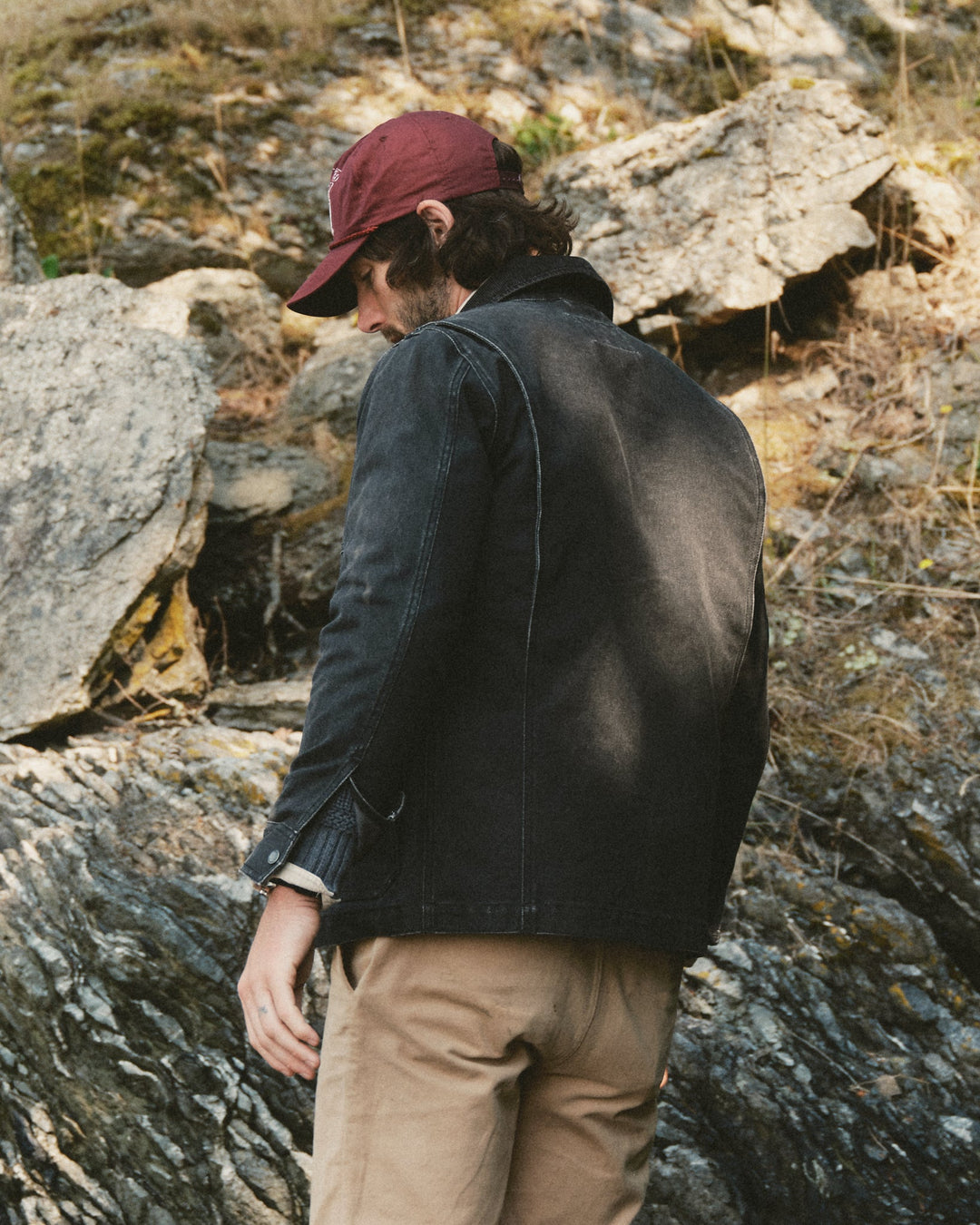 A man wearing a dark charcoal jacket, tan pants, and a maroon baseball cap stands outdoors against a rocky, natural background, viewed from behind.