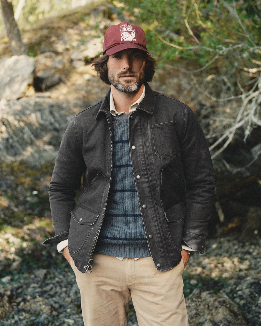 A man wearing a dark charcoal canvas jacket, a blue-gray striped knit sweater, beige pants, and a maroon baseball cap, standing outdoors in a rocky, wooded environment.