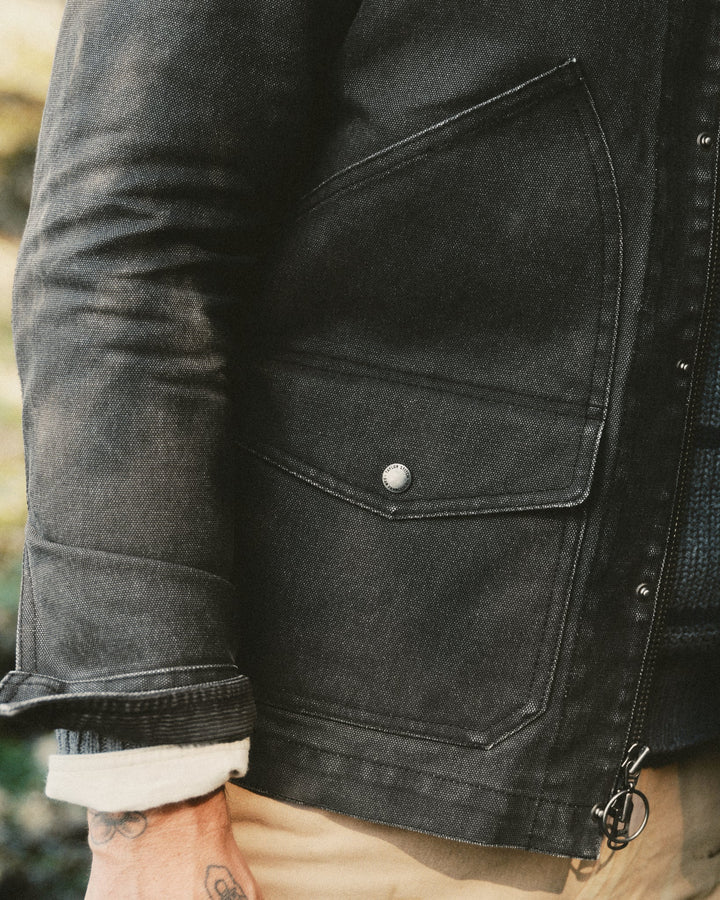 Close-up of a person wearing a dark charcoal gray canvas utility jacket with a snap-button flap pocket, zippered front, and a light-colored shirt cuff visible at the sleeve.